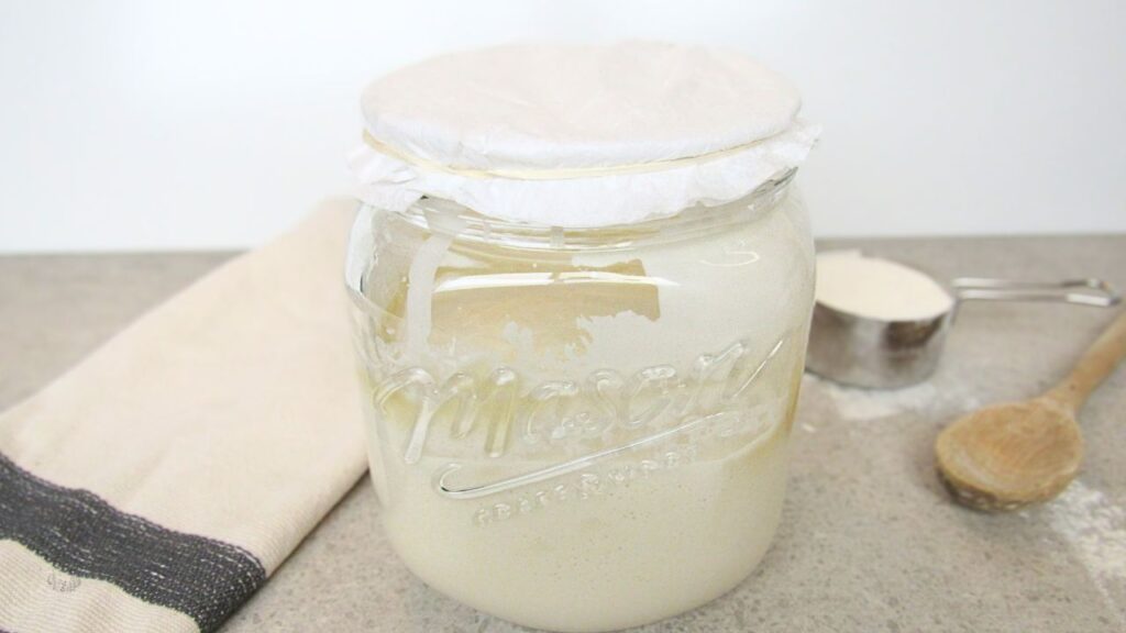 słoik z zakwasem przykryty filtrem do kawy, a w tle łyżka drewniana, kubek mąki pszennej i ścierka