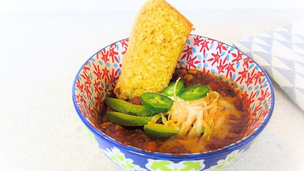 miska z chili con carne przyozdobionym serem, awokado i papryczkami jalapeno, a w chili zanurzony chleb kukurydziany