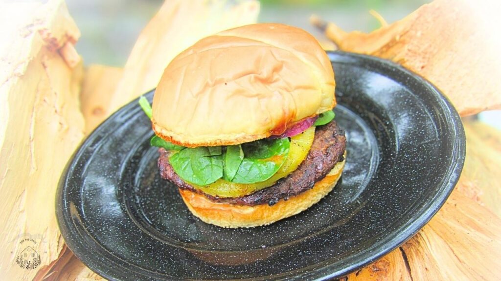 burger wołowy po hawajsku z ananasem na czarnym talerzu metalowym otoczony drewnem na ognisko