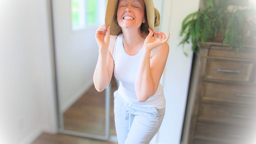 dziewczyna w białej koszulce i szarych spodniach śmiejąca się i trzymająca kapelusz słoneczny